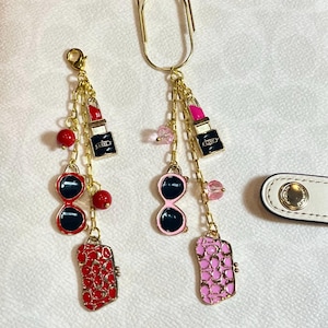 May include: Two gold-toned paper clips with dangling charms. One clip has a lipstick charm, sunglasses, and a red patterned purse charm. The other clip has a lipstick charm, sunglasses, and a pink patterned purse charm. Red and pink accents.