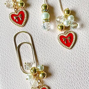 May include: Three gold heart-shaped charms with red enamel and a bow design. Each charm is attached to a string of gold beads, pearls, and clear crystals.