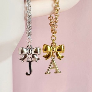 May include: Two gold chain bracelets with a bow charm and a letter pendant. The bracelet on the left has a silver bow and a letter J pendant. The bracelet on the right has a gold bow and a letter A pendant.