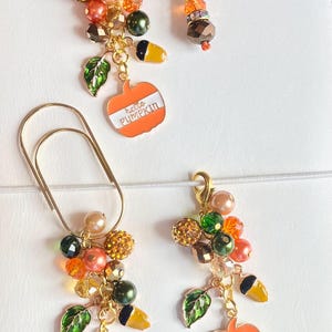 May include: Two gold-tone pumpkin charms with the text "hello pumpkin" on each. Each charm is attached to a string of beads in orange, green, and gold tones. The charms are attached to a gold-tone paper clip and a gold-tone clip.