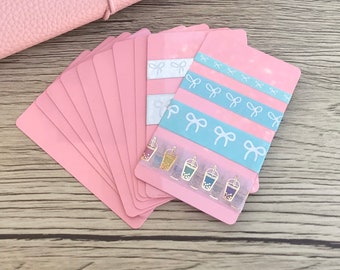 May include: A stack of pink cardstock paper with decorative washi tape featuring a pattern of white bows and gold bubble tea cups on the top card.