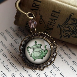 Pode incluir: Um amuleto de vidro com uma moldura de metal bronze que representa um bule de chá verde e branco. O amuleto está preso a um marcador de página de bronze com uma conta rosa.