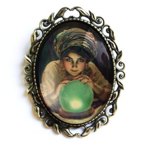 May include: A vintage-style brooch featuring a woman in a turban gazing into a green crystal ball. The brooch is made of metal with an oval-shaped glass cabochon.