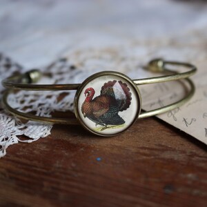 May include: A gold metal bangle bracelet with a round glass charm featuring a vintage illustration of a brown and white turkey.