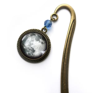 May include: A bronze bookmark featuring a round, glass moon charm and a blue crystal bead. The moon charm depicts a detailed image of the lunar surface.