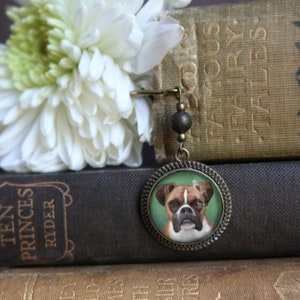 May include: A bronze bookmark with a round glass charm featuring a portrait of a brown and white boxer dog on a green background. The bookmark is hanging from a chain with a brown bead.