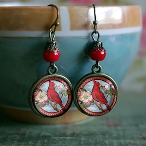 May include: A pair of dangle earrings with red glass beads and round glass cabochons featuring a red cardinal bird perched on a branch with white blossoms. The cabochons are set in bronze-toned metal.