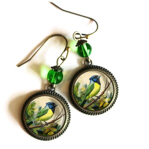 May include: A pair of earrings featuring a green glass bead and a round pendant with a vintage illustration of a blue and yellow bird perched on a branch. The pendant is framed in a bronze-colored metal.
