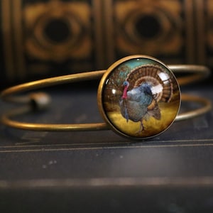 Puede incluir: Un brazalete de brazalete de latón con un encanto de vidrio redondo que presenta una imagen detallada de un pavo con sus plumas extendidas. El pavo está de pie en un campo con un fondo de árboles y cielo.