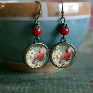 May include: A pair of dangle earrings with red glass beads and round glass charms featuring a cardinal bird perched on a branch with white blossoms. The charms are set in bronze-toned metal.