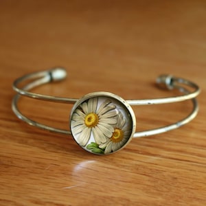 May include: A silver bangle bracelet with a round glass cabochon featuring a pressed white daisy with a yellow centre.
