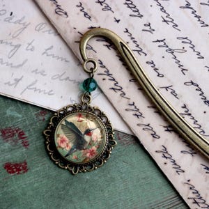 May include: A bronze bookmark with a green glass bead charm and a round, ornate metal pendant featuring a hummingbird in flight against a floral background.