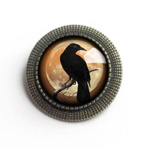 May include: A black raven perched on a branch in front of a large, orange moon. The raven is silhouetted against the moon. The image is framed in a round, antique-looking metal frame.