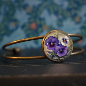 May include: A brass bangle bracelet with a round glass cabochon featuring a hand-painted design of three purple and white pansies.
