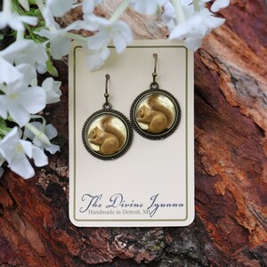 May include: A pair of gold-tone dangle earrings featuring a round, clear glass cabochon with a painted image of a brown squirrel. The earrings are on a white card with the text "The Divine Iguana Handmade in Detroit, MI".