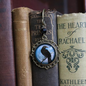 May include: A bronze bookmark with a black raven silhouette on a blue moon background. The bookmark is hanging from a book with the title "The Heart of Rachael" on the spine.
