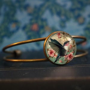 Puede incluir: Un brazalete de brazalete de latón con un cabujón redondo de vidrio que presenta un colibrí en vuelo rodeado de flores rosas sobre un fondo verde.