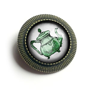 May include: A green teapot with a white background, framed in a bronze-colored metal circle with a textured edge.