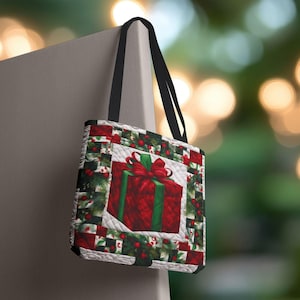 May include: A quilted tote bag featuring a large red gift box with a green bow and ribbon. The bag has a black handle and a festive holiday design with red berries and green foliage. The bag is hanging on a gray surface.