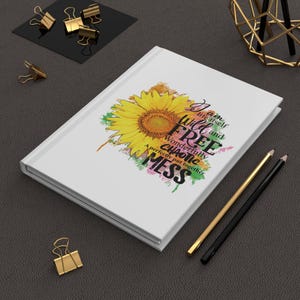 Puede incluir: Diario blanco con un diseño de girasol y el texto "I am life itself, wild and free, wonderfully chaotic mess, a perfectly put together." También se ven lápices dorados y negros y clips dorados.
