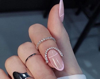 May include: A hand with several rings on the fingers, including a silver ring with a row of small diamonds. The ring is on the ring finger. The nail on the ring finger is painted with a light pink polish and has a white line across the top. The nail is oval shaped.