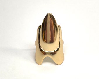 May include: A gold-colored ring shaped like a long, pointed nail. The ring is designed to be worn on the finger, with the pointed end extending upwards.