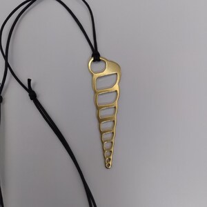 Puede incluir: Un collar con colgante dorado con diseño de concha estilizada. El colgante está suspendido de un cordón negro. El diseño de la concha tiene una serie de segmentos curvos conectados, creando un efecto espiral. El fondo es gris neutro.