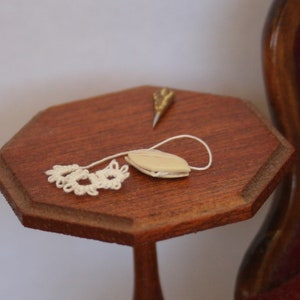 Könnte beinhalten: Ein kleiner, achteckiger Holztisch. Auf dem Tisch liegen ein cremefarbenes Objekt mit weißer Schnur und eine goldfarbene Nadel. Ein cremefarbenes, gehäkeltes Blumendesign ist an der Schnur befestigt.