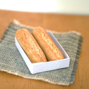 May include: Two miniature, golden-brown baguettes sit in a white rectangular container, placed on a textured, light-green napkin. The bread has a detailed, textured surface, suggesting a fresh bake. The scene is set on a wooden surface.