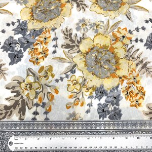 K&ouml;nnte beinhalten: Wei&szlig;er Stoff mit einem floralen Muster mit gelben, orangefarbenen und grauen Blumen. Der Stoff hat einen grauen Rand mit einem sich wiederholenden Muster.