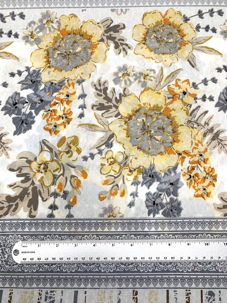 K&ouml;nnte beinhalten: Wei&szlig;er Stoff mit einem floralen Muster mit gelben, orangefarbenen und grauen Blumen. Der Stoff hat einen grauen Rand mit einem sich wiederholenden Muster.