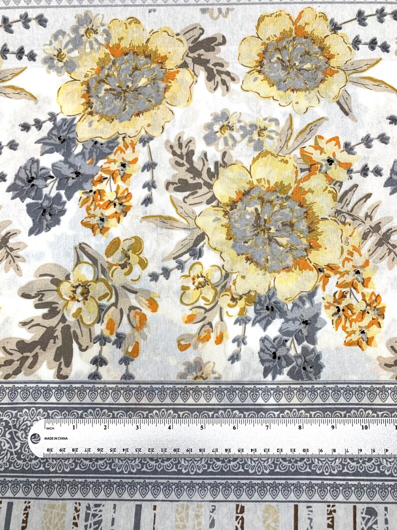 K&ouml;nnte beinhalten: Wei&szlig;er Stoff mit einem floralen Muster mit gelben, orangefarbenen und grauen Blumen. Der Stoff hat einen grauen Rand mit einem sich wiederholenden Muster.