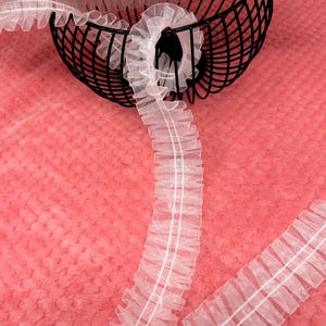May include: White ruffled ribbon with a central white stripe, displayed against a pink textured background. The ribbon is partially coiled in a black wire basket. The delicate fabric suggests use in crafts or sewing projects.