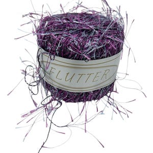 May include: A ball of purple and silver yarn with the word "FLUTTER" on a label. The yarn has a textured appearance with strands extending outward. Ideal for knitting and crochet projects.