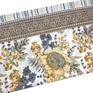 K&ouml;nnte beinhalten: Wei&szlig;er Stoff mit einem floralen Muster in Graut&ouml;nen, Gelb und Braun. Der Stoff hat einen Rand mit einem sich wiederholenden Muster.