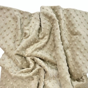May include: A beige blanket featuring a raised dimpled texture across the entire surface. The fabric has a soft, plush appearance with a consistent light tan color and a repeating pattern of small, rounded bumps throughout the material.