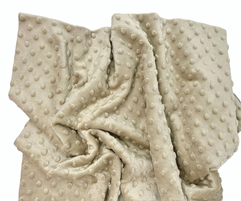 May include: A beige blanket featuring a raised dimpled texture across the entire surface. The fabric has a soft, plush appearance with a consistent light tan color and a repeating pattern of small, rounded bumps throughout the material.