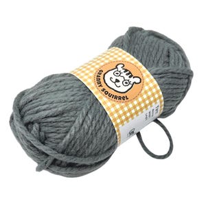 May include: A skein of gray yarn with a label featuring a cartoon squirrel wearing glasses. The label has a yellow and white checkered pattern and the text "Granny Squirrel". The yarn is thick and appears soft, ideal for knitting or crocheting projects.