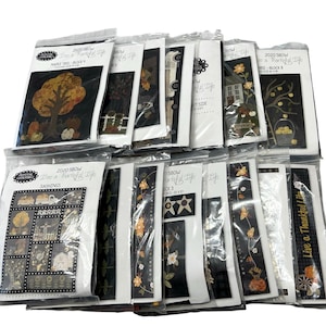 May include: A collection of twelve individual quilt block patterns from the 2020 Live a Thankful Life series by Primitive Gatherings, each sealed in a clear plastic sleeve featuring printed illustrations of seasonal motifs like trees, pumpkins, and houses.