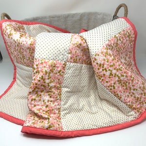 May include: A pink and white patchwork baby quilt with gold polka dots and confetti print. The quilt is folded and placed in a woven basket.