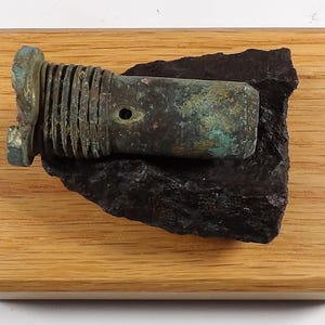 May include: An antique bronze object with a threaded section and a cylindrical body, displaying a green patina. It rests on a piece of dark, rough stone, mounted on a rectangular wooden base. The object has a small hole.