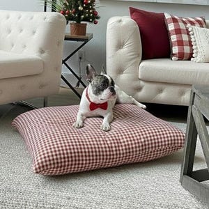 May include: A French Bulldog wearing a red bow tie lies on a red and white checked dog bed. The dog bed is a square cushion. The dog is in a living room with a Christmas tree and a white sofa.