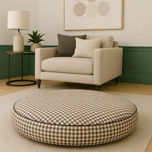 May include: A round, brown and cream checkered pet bed sits on a light beige rug. A beige armchair with two throw pillows is in the background. A small side table with a lamp and a potted plant are to the left.
