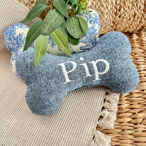 May include: A blue dog bone-shaped toy with the name "Pip" embroidered in white. The toy has a herringbone pattern. A blue and white floral patterned pillow is behind the toy, with green leaves on top. The toy is on a beige surface.