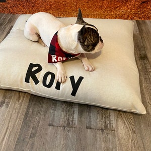 May include: A white and brown French bulldog wearing a red and black bandana lies on a beige dog bed with the name "Roy" embroidered on it.