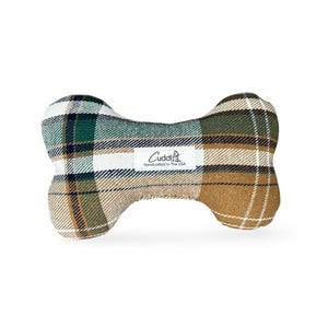 Könnte beinhalten: Ein knochenförmiges Hundespielzeug mit einem karierten Muster in Grün, Weiß und Braun. Das Spielzeug hat ein weißes Etikett mit dem Wort "Cuddli" und "Handcrafted In The USA".