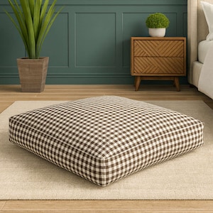 Può includere: Un cuscino da pavimento quadrato con un motivo a quadri marrone e bianco. Il cuscino è posizionato su un tappeto beige chiaro. Lo sfondo include una parete verde, un comodino in legno e una pianta in vaso.