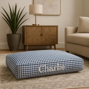 May include: A square dog bed with a blue and white gingham pattern. The word "Charlie" is embroidered in white on the front of the bed. The bed is placed on a neutral-colored rug in a living room setting.