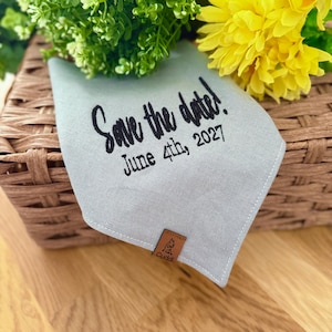 May include: A light blue dog bandana with the text "Save the date! June 4th, 2027" in black script. The bandana has a small brown leather tag with a dog silhouette and the word "Cuddl".