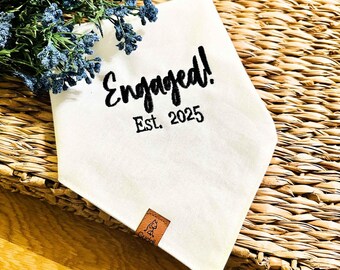 Peut inclure: Un bandana blanc pour chien avec le mot "Engaged!" et "Est. 2025" imprimé en noir. Une petite étiquette en cuir marron avec une illustration de chien est attachée. Des fleurs bleues sont dans le coin supérieur gauche.